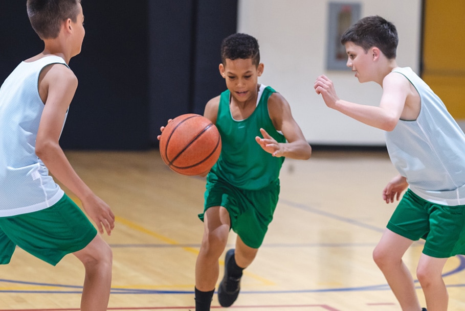 A boy in a green jersey dribbles a basketball between two opposing players.